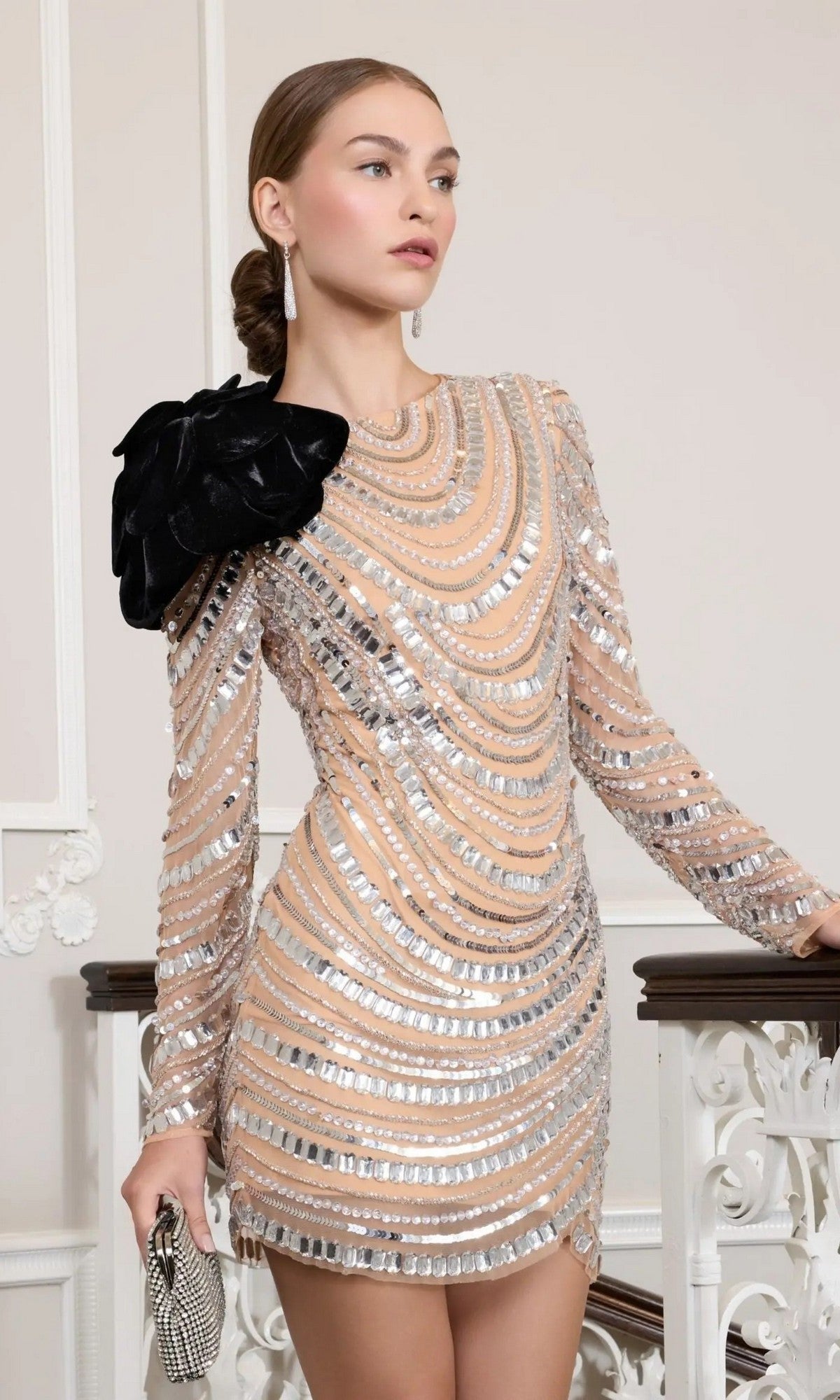 Woman wearing the Mac Duggal 94284 short party dress in Silver Nude, featuring sparkling sequins and silver patterns with a statement black flower on the shoulder. She holds a small clutch and stands beside a decorative white railing.